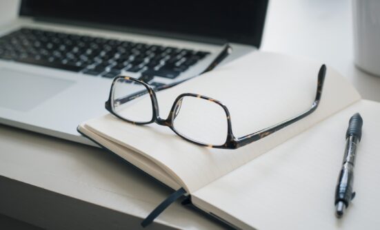 Glasses on a notebook, next to a pen and laptop