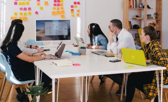 A group of people sitting around a table with laptops, looking at a large monitor