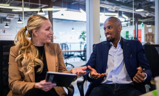 A man and women at work discussing something and referring to a notebook