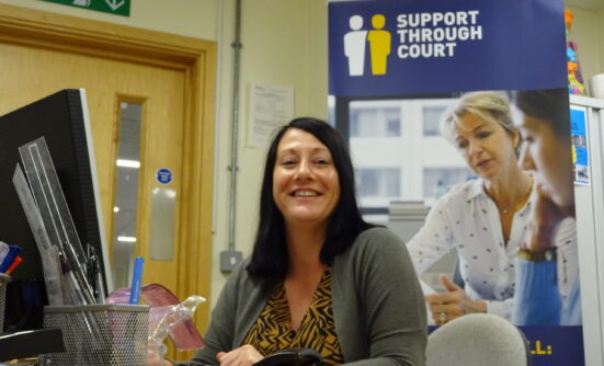 Support Through Court Worker sitting at her desk and smiling, with a poster of 'Support Through Court' in the background