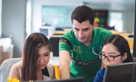 a man leaning over two women working on their computers, showing them something.